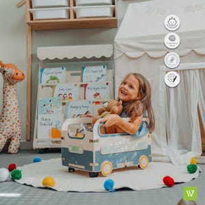 Puede incluir: Una caja de juguetes azul y blanca con forma de furgoneta vintage con ruedas amarillas. Una ni&ntilde;a est&aacute; sentada en la caja de juguetes, sosteniendo un animal de peluche. La caja de juguetes est&aacute; sobre una alfombra blanca con pompones de colores.