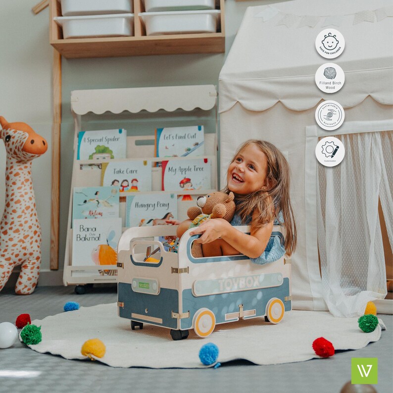 Puede incluir: Una caja de juguetes azul y blanca con forma de furgoneta vintage con ruedas amarillas. Una ni&ntilde;a est&aacute; sentada en la caja de juguetes, sosteniendo un animal de peluche. La caja de juguetes est&aacute; sobre una alfombra blanca con pompones de colores.
