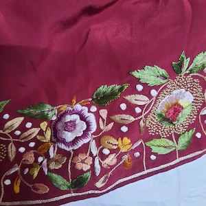 Pode incluir: Close-up de um tecido bordô com uma borda floral bordada. O bordado apresenta flores e folhas brancas, roxas, amarelas e verdes. O design é detalhado com um contorno dourado e pontos brancos.