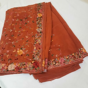 Peut inclure: Textile de couleur rouille avec broderies florales complexes. La broderie présente diverses fleurs dans les tons de jaune, rose et rouge, ainsi que des feuilles et des vignes vertes. Le tissu est plié, mettant en valeur le motif de la bordure.