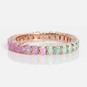May include: Rose gold ring featuring a band of colorful opal stones. The opal stones transition in color from pink to purple to blue-green. The ring is set against a white background, with a reflection visible below.