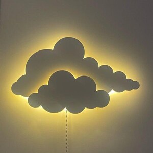 May include: A white cloud-shaped wall light with a warm yellow glow. The light is attached to the wall with a cord.