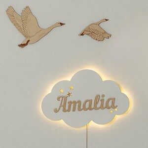 May include: A white wooden cloud-shaped night light with the name "Amalia" written in a cursive font. The light is illuminated and has small gold stars cut out around the name. Two wooden geese are mounted on the wall behind the light.