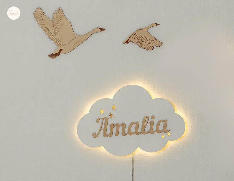 May include: A white wooden cloud-shaped night light with the name "Amalia" written in a cursive font. The light is illuminated and has small gold stars cut out around the name. Two wooden geese are mounted on the wall behind the light.