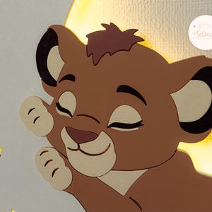 May include: A wooden wall decoration featuring a cartoon lion cub with closed eyes, a light brown mane, and a light beige face. The lion cub is set against a yellow crescent moon and surrounded by yellow star cutouts. The Telman logo is in the upper right corner.