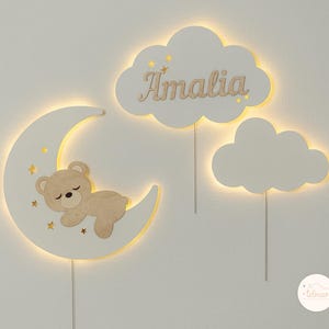 May include: A white wooden moon with a sleeping teddy bear inside, two white clouds with the name "Amalia" on one, and a small white cloud with the word "telman" on it.