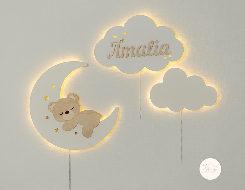 May include: A white wooden moon with a sleeping teddy bear inside, two white clouds with the name "Amalia" on one, and a small white cloud with the word "telman" on it.
