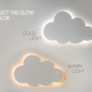 May include: Two cloud-shaped wall lights with illuminated edges are displayed. The top cloud emits a gold light, and the bottom cloud emits a warm light. Text above reads "SELECT THE GLOW COLOR". Text next to the clouds reads "GOLD LIGHT" and "WARM LIGHT".