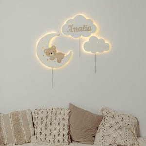 May include: A white wooden moon with a sleeping bear and stars, two white wooden clouds, one with the name "Amalia" on it. The moon and clouds are lit up with warm white lights. The wall decor is hanging on a white wall above a couch with beige pillows.