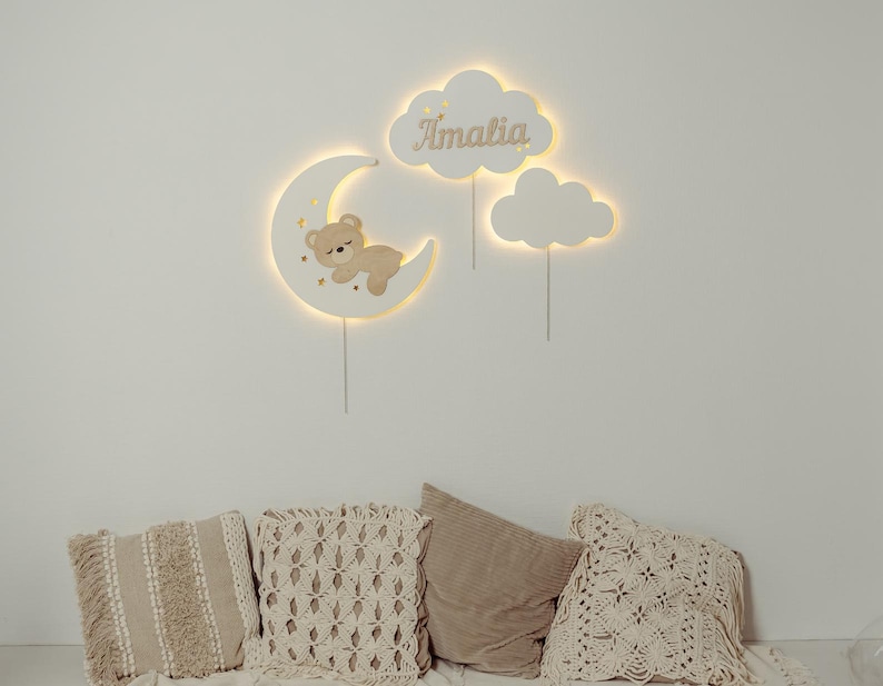 May include: A white wooden moon with a sleeping bear and stars, two white wooden clouds, one with the name "Amalia" on it. The moon and clouds are lit up with warm white lights. The wall decor is hanging on a white wall above a couch with beige pillows.