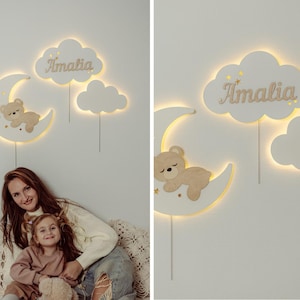 May include: A white wooden nightlight with a crescent moon, a cloud with the name "Amalia" and a cloud with a sleeping bear. The nightlight is illuminated with warm white LED lights.