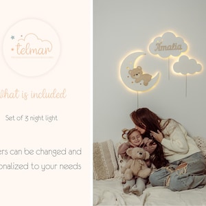 May include: A set of three white wooden night lights shaped like clouds and a crescent moon. The moon features a small brown teddy bear. The cloud with the name "Amalia" is lit up.