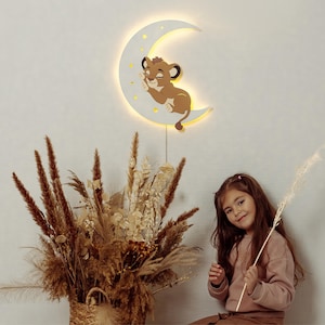 May include: A wooden crescent moon night light with a sleeping lion cub design. The light is warm and inviting, perfect for a child's room. The light is hanging on a wall.