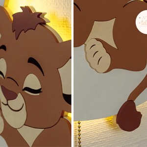 May include: A brown and beige lion cub-shaped wall lamp. The lamp features a smiling lion cub with closed eyes and a tail. The lamp is made of wood and has a warm, yellow light. The lamp is designed for a nursery or children's room.