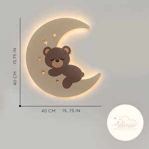 May include: A beige wooden crescent moon night light with a brown bear sleeping on it. The moon has cut-out stars and a warm white glow. The light measures 15.75 inches in diameter.