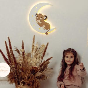 May include: A wooden night light shaped like a crescent moon with a sleeping lion cub design. The moon is white with yellow stars and a warm glow. The image also shows a young girl holding a plant.