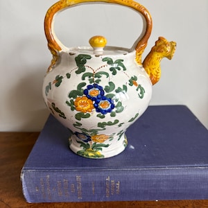 Può includere: Una teiera in ceramica bianca con manico e beccuccio gialli. La teiera è decorata con motivi floreali blu, gialli e verdi. Il beccuccio ha la forma di una testa di drago. La teiera è appoggiata su un libro blu.