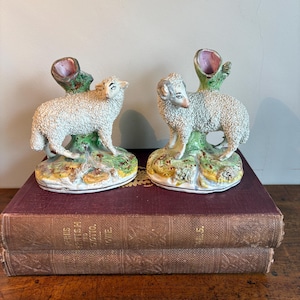 May include: A pair of ceramic sheep figurines, each featuring a textured white wool coat, a green tree stump with a pink-lined opening, and a painted base. The sheep are standing on a grassy, multi-coloured ground with detailed facial features.