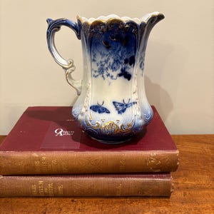 May include: Vintage porcelain jug with a blue and white floral and butterfly pattern. The jug features a curved handle and gold detailing on the rim and base. It rests on two antique books with red covers.
