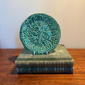 May include: A green ceramic plate with a textured leaf design, displayed on a white stand. The plate is placed on top of two antique books with green covers. The books are stacked on a wooden surface.