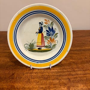 Puede incluir: Un plato de cerámica decorativo con un borde amarillo y azul. El plato presenta una imagen pintada a mano de una mujer con vestimenta tradicional, de pie cerca de flores estilizadas. El plato se exhibe sobre una superficie de madera.