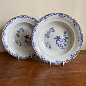 Könnte beinhalten: Zwei weiße Suppenschalen aus Keramik mit einem blauen und goldenen Blumendesign. Die Schalen haben einen gewellten Rand und einen goldenen Rand.