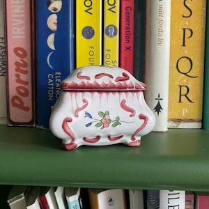 Könnte beinhalten: Eine weiße Keramik-Schmuckschatulle mit einem rosafarbenen Deckel und einem Blumendesign. Die Schachtel hat einen gewellten Rand und vier kleine Füße. Die Schachtel steht auf einem grünen Regal mit Büchern dahinter.