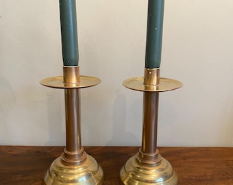 May include: A pair of brass candle holders with a simple design. Each holder has a round base, a tall cylindrical stem, and a small round plate for holding the candle. The candles are dark green.