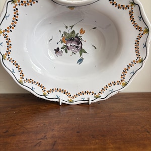 May include: An oval ceramic bowl with a scalloped rim, featuring a white base with a hand-painted floral motif in the center. The border is decorated with a repeating pattern of small orange and black dots and blue accents along the edge.