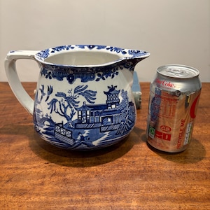 May include: A blue and white ceramic pitcher with a handle, featuring a Willow pattern design. The pitcher is next to a Coca-Cola can. The pitcher is a vintage item.