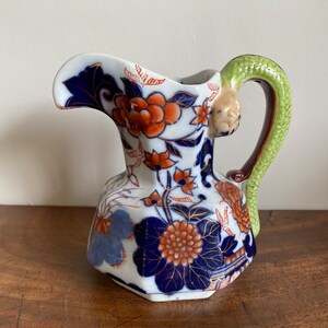 May include: A small, white ceramic jug with a blue and orange floral pattern. The jug has a green and brown snake-like handle.