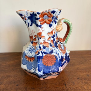 Peut inclure: Un pichet en céramique à base blanche orné d'un motif floral aux tons orange et bleu profond. Le récipient présente un corps hexagonal, un bord festonné et une anse verte texturée avec un détail sculpté au point de fixation supérieur.
