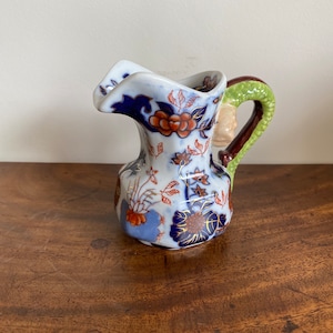 Peut inclure: Un petit pichet en porcelaine bleu et blanc avec une anse verte et brune. Le pichet est décoré d'un motif floral dans les tons bleus, rouges et dorés.