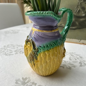 May include: A yellow, green, and purple ceramic pitcher with a textured surface and a handle. The pitcher has a floral design and is decorated with a face.