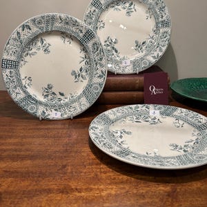 May include: Three antique white porcelain plates with a green floral pattern. The plates have a raised rim and a scalloped edge. The plates are on a wooden table.