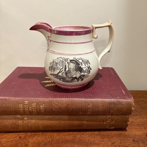 May include: A white ceramic pitcher with a pink rim and handle. The pitcher features a black and white illustration of a woman under a weeping willow tree. The pitcher is sitting on top of two antique books with maroon covers.