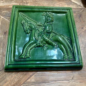 Könnte beinhalten: Quadratische, grüne Keramikfliese mit einem Relief einer Person auf einem Pferd. Der Reiter trägt einen Hut und hält einen Speer. Das Pferd ist in Bewegung. Die Fliese hat eine glänzende Oberfläche und einen erhabenen Rand.