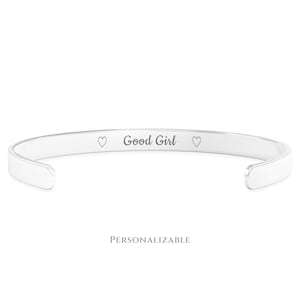 May include: A silver-colored cuff bracelet with the words "Good Girl" engraved in the center, flanked by two heart symbols. The word "PERSONALIZABLE" is printed below the bracelet.
