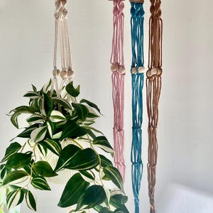 Può includere: Tre portavasi in macramè con perline di legno. I portavasi sono di colore bianco, rosa e marrone. Il portavasi bianco ha una pianta dentro.