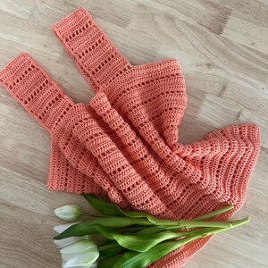May include: A coral-colored crocheted tank top with a delicate open-stitch pattern. The top is laid flat on a wooden surface with a bouquet of white tulips.
