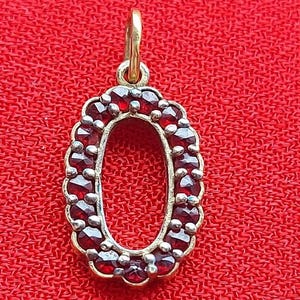 May include: A gold-coloured pendant shaped like the letter "O", adorned with small, round, deep red gemstones. The pendant has a loop at the top for a chain and is set against a textured red background. The gemstones are closely set.