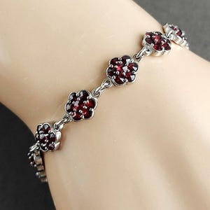 Op de afbeelding: Een zilverkleurige schakelarmband met een reeks bloemvormige zettingen. Elke bloem bestaat uit meerdere dieprode, gefacetteerde stenen die in een cluster zijn gerangschikt. De armband is opgebouwd uit scharnierende metalen schakels tussen de bloemen.