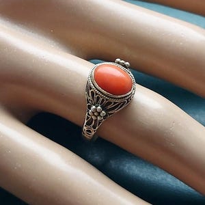 May include: A gold-toned ring featuring an oval, coral-colored stone. The stone is set within a filigree design with floral accents. The ring's band has an intricate, openwork pattern. The ring is displayed on a light-toned surface.