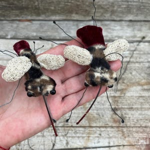 Könnte beinhalten: Zwei insektenförmige Ornamente mit dunkelroten Samtköpfen, flauschigen braun-schwarzen Körpern und strukturierten cremeweißen Netzflügeln. Jedes Ornament hat dünne schwarze Drahtantennen und Beine sowie kleine schwarze Perlenaugen an der Vorderseite.