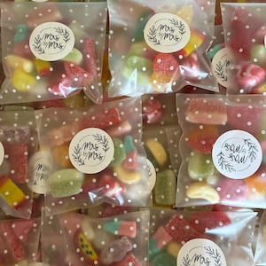 May include: Close-up of several small cellophane bags filled with assorted gummy sweets. Each bag is sealed with a white sticker that reads "Mrs & Mrs" in a decorative font, surrounded by a wreath. The bags are decorated with white polka dots.