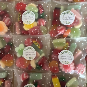 May include: Several cellophane bags filled with assorted gummy sweets. Each bag is sealed with a circular sticker that reads "Love is Sweet Enjoy this Treat". The sweets are in various shapes and colours, including red, green, yellow, and pink.