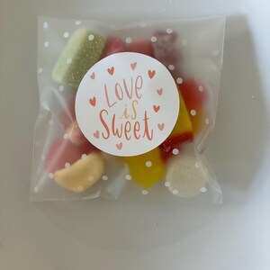 May include: A square, translucent plastic pouch with a white polka dot pattern containing assorted colourful gummy sweets. A circular white sticker is centred on the front, featuring the text "Love is Sweet" in orange script surrounded by small red hearts.