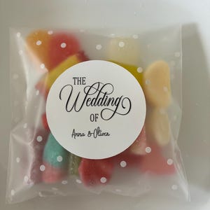 May include: A translucent plastic bag with a white polka dot pattern containing assorted colourful gummy sweets. A circular white sticker is centred on the front, featuring the black text "THE Wedding OF Anna & Oliver" in a decorative script font.
