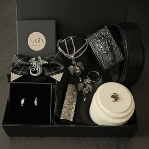 Peut inclure: Un coffret noir contenant divers accessoires sur le thème du dragon. L'ensemble comprend un nœud papillon, un collier, des boucles d'oreilles, une ceinture, un briquet, un porte-clés et un petit récipient rond. Les accessoires sont argentés. Le coffret porte le texte "NAJIA".