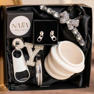 Puede incluir: Una caja de regalo con varios accesorios, incluyendo pendientes plateados con forma de cremallera, un abrebotellas, una pulsera y una pinza para el pelo adornada con pedrería. La caja también contiene un objeto de cerámica blanca y una letra "Y" adornada con pedrería. La caja está forrada con satén negro.